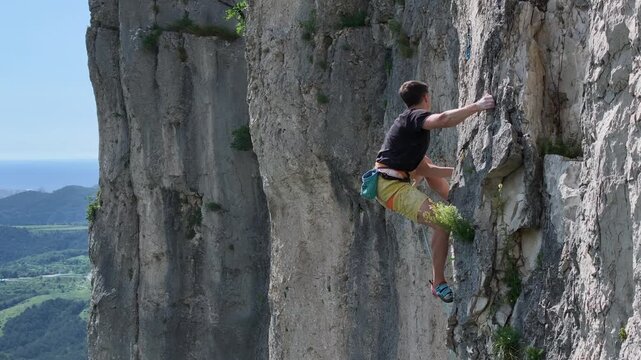 AERIAL: Male climber scales a vertical rock face with stunning view of the Adriatic Sea and distant hills. Young man training in a picturesque sport climbing area surrounded by the Karst landscape.