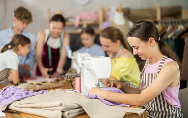 Teenage girl working at sewing machine during class in cozy school workshop while female teacher helping classmates grasp basics of tailoring