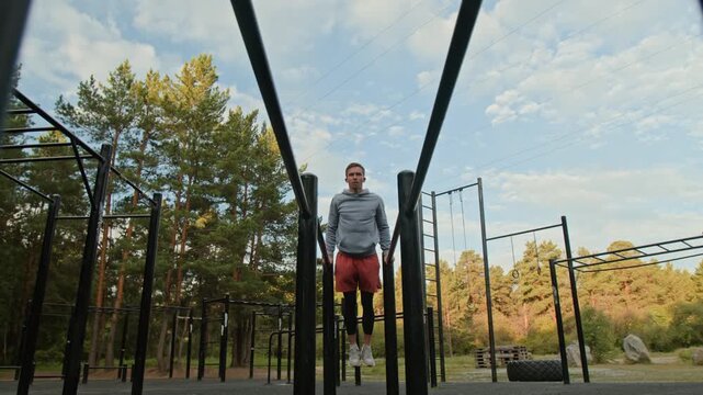 Low angle shot of young man in sportswear performing dips on parallel bars amidst trees in outoor gym