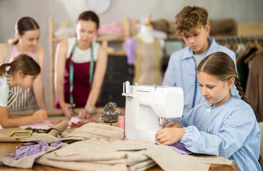 Focused and diligent ten year old girl carefully sewing on machine during hands-on class, while classmates preparing patterns and fabric under guidance of female teacher