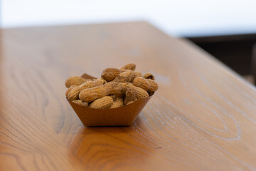 A paper basket containing dried salted peanuts on a wooden table. The salty snacks are whole with the shells dried. The tubular-shaped protein is tan colored with a papery texture on the outside nut.