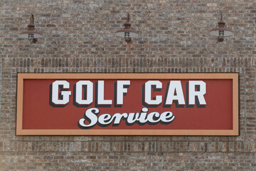 Fototapeta premium A red and yellow golf car service sign with white text and black trim. The sign is affixed to a brown and beige colored brick building with three barn lights over the signage for mechanical servicing.
