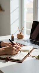 Writing Inspiration: Hands of an anonymous person diligently write in a notebook amidst a modern office setting, the presence of a laptop and fragrance bottle suggesting a comfortable.