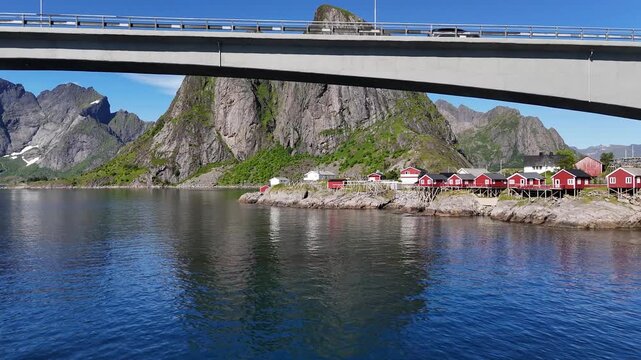 Drone flying under the car bridge and over small fishing village Hamn&oslash;y in Norway towards iconic mountains above the sea with wooden drying racks for torrfisk