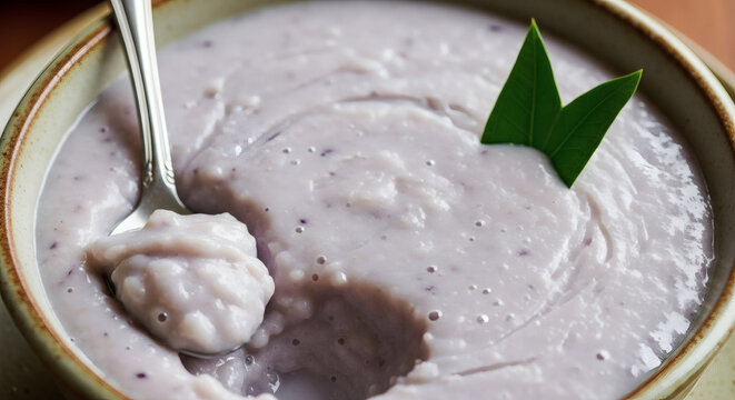 Close up of a bowl of bubur pulut hitam with a spoon and a pandan leaf garnish on top of the dessert