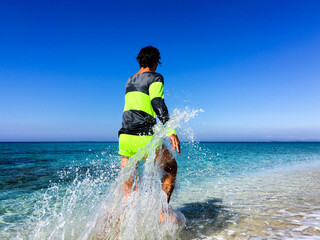Sporty man in neon sportswear running back to the camera on the beach next to turquoise coloured...