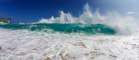 Dramatic turquoise wave crashing onto a sandy beach under a
clear blue sky, with foamy water and dynamic sea spray.