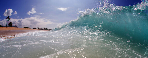 Dramatic turquoise wave crashing onto a sandy beach under a clear blue sky, with foamy water and...