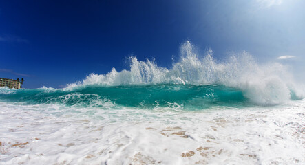 Obraz premium Dramatic turquoise wave crashing onto a sandy beach under a clear blue sky, with foamy water and dynamic sea spray.