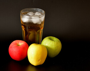 A mixture of apple juices of different varieties in a tall faceted glass with ice on a black background, next to ripe fruit.