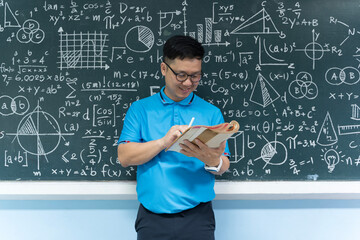 Mathematics teacher reading book and writing notes in front of blackboard with equations in classroom