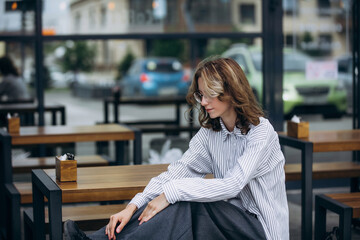 A teenage girl model is young and beautiful in a shirt and trousers on the street in a street cafe
