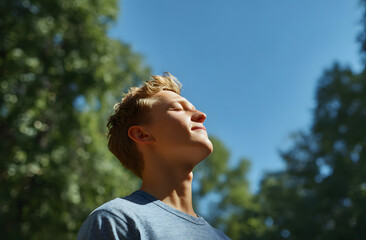 Young man breathes fresh air in natural atmosphere. Adult guy tilts head up and closes eyes in forest and blue sky. He enjoys vacation, meditates, feels calm.