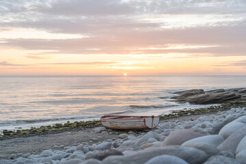 Tranquil coastal sunrise scene featuring an old boat on a pebble beach near the ocean