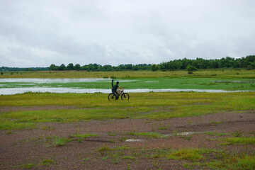 A man rides a bicycle across a wide green field with wetlands and trees in the background in a rural area