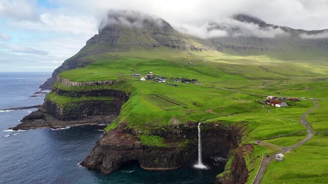 Aerial view of G&aacute;sadalur waterfall plunging into the Atlantic Ocean, framed by dramatic cliffs, lush green Faroe Islands landscape, and traditional village houses
