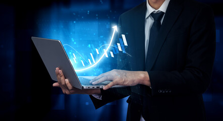 A businessman in a suit is engaged with a laptop in a modern office, showcasing digital graphs and upward trends that indicate growth and success. Impute
