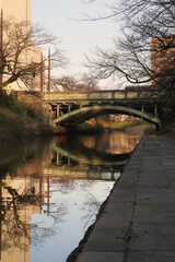 Obraz premium Peaceful riverside walkway in Autumn. A paved path runs alongside a calm river with a bridge and bare trees reflected in the water on a tranquil autumn day.