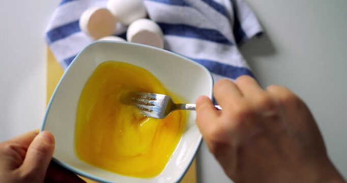 Closeup of hand whisking egg mixture in white bowl with fork, striped kitchen towel and egg shells nearby, top view breakfast preparation.