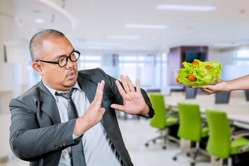 Disgusted man refusing a salad. A business man with a disgusted and shocked expression is holding a hand up to stop someone from offering a bowl of salad, with the person's hand visible on the side.