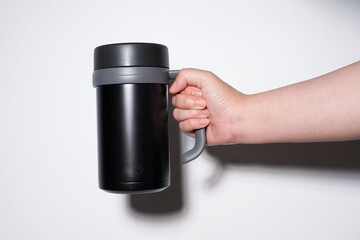Hand holding simple black colored thermos mug water bottle with handle isolated on horizontal white background.