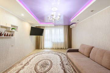 Cozy living room with a beige sofa, oval carpet, TV, and a chandelier under a purple-lit ceiling