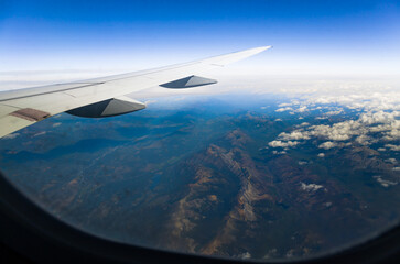 View through an airplane window catching the amazing mountains through  the clouds, with the plane's wing prominently displayed against the horizon.