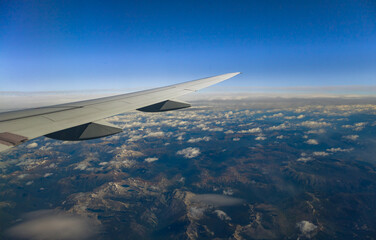 View through an airplane window catching the amazing mountains through  the clouds, with the plane's wing prominently displayed against the horizon.