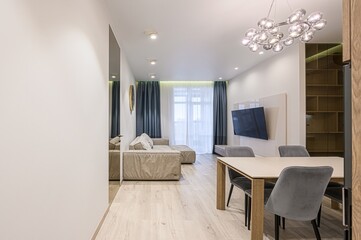 modern, bright living room with a light-colored sofa, dining table, and a large TV. Features include hardwood floors, blue curtains, and a stylish chandelier