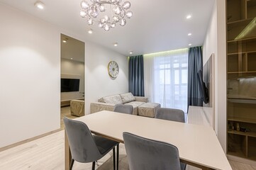 stylish living room featuring a dining table with gray chairs, a beige sofa, and a modern chandelier. The room is bright and well-lit, with a cozy and inviting atmosphere