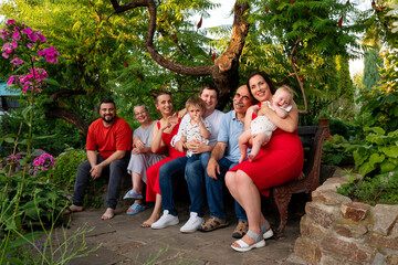 A close-knit family shares a warm, affectionate moment seated together on a garden bench, surrounded by lush greenery and vivid blossoms on a sunny summer day