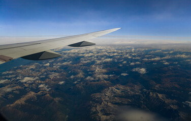 View through an airplane window catching the amazing mountains through  the clouds, with the plane's wing prominently displayed against the horizon.