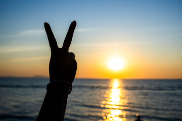 silhouetted hand making a peace sign against a glowing sunset over the ocean, with golden light
