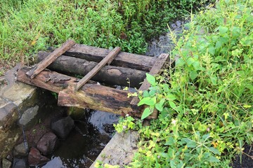 Obraz premium A simple rustic wooden bridge made from old logs and planks placed across a small stream in the countryside. Surrounded by lush green grass, wild plants, and natural stones.