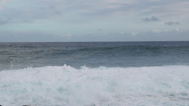 A rising set collides with a reef rock and erupts into white spray, churning foam across the frame while gray skies and restless swell convey raw, wind-driven energy.