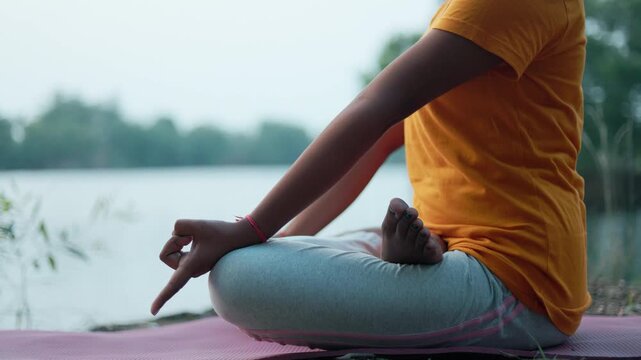 Crop view of a woman sitting in padmasana pose with apana mudra near a lake during blue hour twilight.