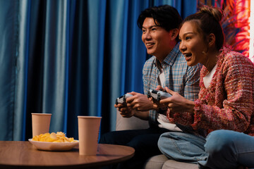 Sitting sofa of happy smiling couple playing video game control by joystick fronted snack and drinks competing challenge level with funny at modern neon blub light comfy living room at home. Infobahn.
