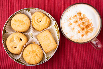 Top view of assorted butter cookies in a classic tin with a frothy cappuccino sprinkled with cinnamon on red background. Perfect for cozy, holiday, or coffee break concepts.