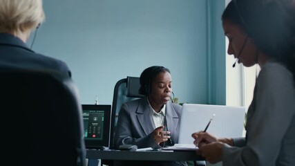 Focus on young African American female manager of call center speaking in headset microphone and looking at laptop screen while consulting online consumers between two diverse colleagues - Powered by Adobe