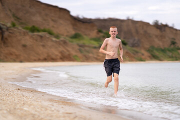 Teenage boy running along the seashore on a warm day. Cheerful guy enjoying a cloudy morning on the seashore. Happy teenager spending summer holidays on a sandy beach.