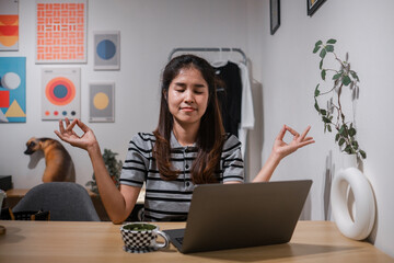 A serene woman meditates at her home office desk, surrounded by modern art and a laptop, embodying calm and focus.