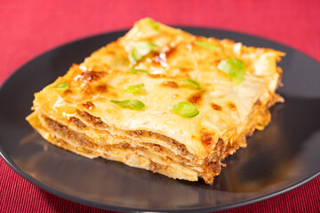 Close-up of a hot lasagna portion with golden melted cheese and basil leaves, served on a black plate. Perfect for Italian cuisine, restaurant menus, and comfort food themes.