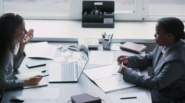Side view of young diverse female office workers sitting in front of each other by desk while one of them describing curious working case to colleague in headset