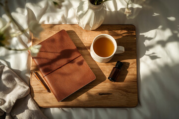 Cozy morning setup with leather notebook, pen, cup of tea, and essential oil bottle on wooden tray in sunlight, symbolizing journaling, mindfulness, relaxation, and self care routine