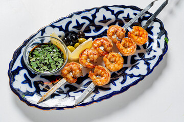 Grilled shrimp skewers resting on a decorative platter beside olives and fresh herbs
