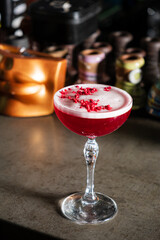 A bright red cocktail with a raspberry sits elegantly on the bar, reflecting ambient light