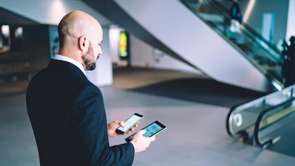 Businessman Examining Mobile Device Near Escalator in Modern Building's Interior - Powered by Adobe