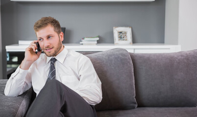 Man in business attire calling on smartphone, sitting on grey sofa near white console, copy space