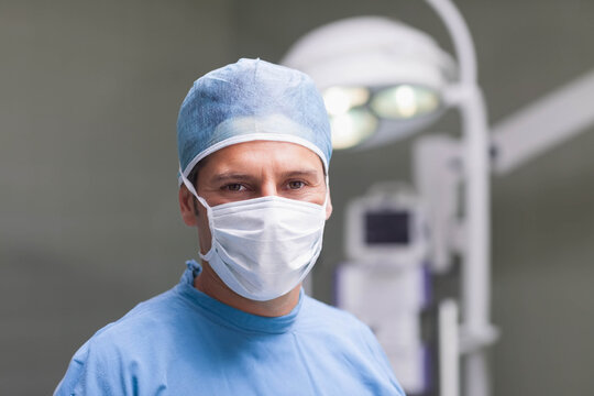 Mid adult male surgeon standing in operating room wearing blue scrubs and mask under overhead lamp