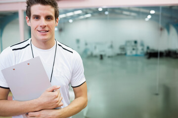 Male coach holding clipboard and wearing whistle standing in gym by glass partition, copy space